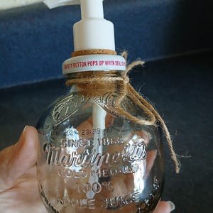 Puede incluir: Una botella de vidrio transparente reutilizada como dispensador de jabón con un mecanismo de bomba blanco. La botella presenta texto en relieve que dice Martinelli's Gold Medal 100% Apple Juice Since 1868. Un trozo de cuerda está atado alrededor del cuello y un anillo de corcho se asienta debajo de la bomba.