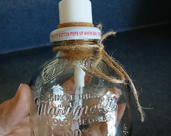 Puede incluir: Una botella de vidrio transparente reutilizada como dispensador de jabón con un mecanismo de bomba blanco. La botella presenta texto en relieve que dice Martinelli's Gold Medal 100% Apple Juice Since 1868. Un trozo de cuerda está atado alrededor del cuello y un anillo de corcho se asienta debajo de la bomba.