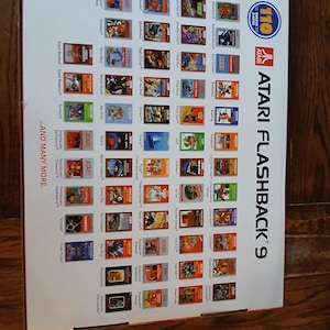 Puede incluir: Una caja blanca de consola de juegos Atari Flashback 9 que presenta una cuadr&iacute;cula de 110 t&iacute;tulos de juegos cl&aacute;sicos, el logotipo de Atari y el texto 110 Built-in Games y ...And Many More. El empaque muestra varias ilustraciones.