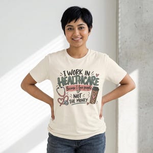 May include: A cream-colored short-sleeve t-shirt featuring the text I WORK IN HEALTHCARE Because I love people NOT THE MONEY. The design includes a pink stethoscope, a leopard print coffee cup with a heart, and small decorative hearts.