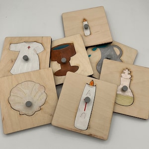 May include: A set of eight wooden puzzle pieces featuring various religious symbols, including a baptismal gown, a chalice, a shell, a candle, a pitcher, and a bottle of oil. The pieces are painted in various colors and have a light wood background.