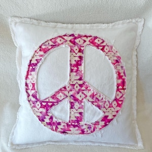Puede incluir: Cojín cuadrado blanco con un gran símbolo de la paz en el centro. El símbolo de la paz es una vibrante mezcla de patrones geométricos rosas y blancos. El cojín tiene un borde con flecos y el texto "BeachRebel.etsy.com" es visible.