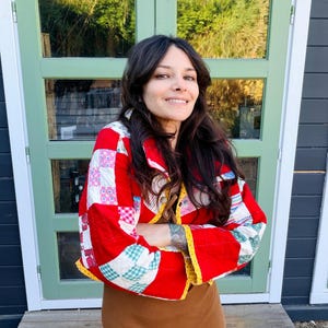 May include: A cropped jacket constructed from a patchwork quilt featuring a vibrant red, white, and multi-colored checkered pattern. The garment has long sleeves, a collar, and a yellow-patterned trim along the edges, worn over a solid brown dress.