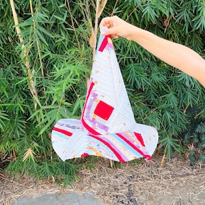 May include: A conical hat constructed from quilted fabric featuring a patchwork design. The surface displays a variety of colors including white, red, blue, and yellow in geometric patterns and stripes, with visible stitching throughout the entire piece.