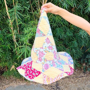 May include: A quilted witch hat featuring a patchwork design of various floral and geometric patterns in shades of yellow, pink, red, and blue. The hat has a tall, pointed crown and a wide, circular brim, constructed from stitched fabric panels.