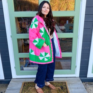 May include: A quilted jacket in bright pink and green with white starburst patterns. The jacket has a relaxed fit and features pockets. The person is wearing dark blue jeans and brown woven shoes. The background includes a green door and a brown doormat.