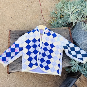 Peut inclure: Une veste matelassée présentant un motif patchwork de carrés bleus, bleu clair et blancs. Le vêtement possède des manches longues, une ouverture frontale aux bords arrondis et un liseré clair le long du col et de l'ourlet, avec une doublure violet clair.