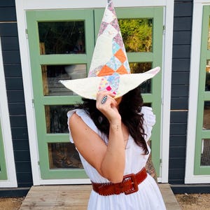 May include: A tall, pointed hat constructed from quilted fabric featuring a patchwork pattern of cream, orange, and floral-print squares. The hat has a wide, slightly curved brim and a visible stitched texture throughout the fabric surface.