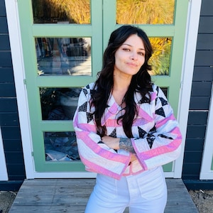 May include: A woman wearing a quilted jacket with a geometric pattern of black, white, and pink triangles and stripes. She is also wearing white pants. The background features green doors and a wooden porch.