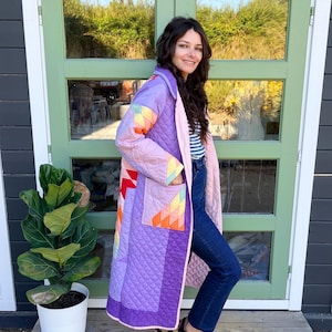 May include: A long, quilted coat in shades of lavender and purple, featuring a geometric patchwork pattern in orange, yellow, and teal on the sleeves and pockets. The coat is worn over a blue and white striped shirt, dark blue jeans, and black clogs.