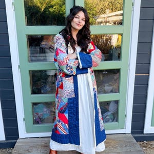 May include: A long, open-front coat featuring a patchwork design with various floral and geometric patterns in red, blue, and white. The coat has solid navy blue trim along the edges and cuffs, worn over a plain white floor-length dress and brown boots.