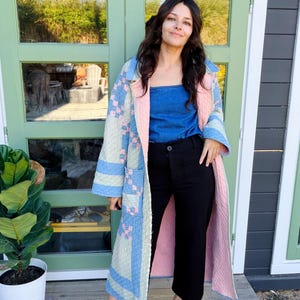 Pode incluir: Um casaco comprido acolchoado com um padrão de patchwork em tons de azul claro, rosa e creme. O casaco tem um forro rosa sólido, mangas compridas e dois bolsos frontais. É usado sobre um top de ganga azul e calças pretas com mules verdes.