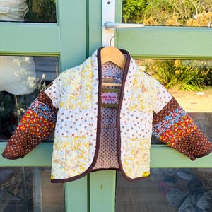 Peut inclure: Une veste matelassée au motif patchwork composé de divers imprimés floraux dans des tons jaune, blanc, rouge et marron. La veste présente des manches longues, un col châle et un liseré marron foncé. Une étiquette indique Milk Thistle Sewing.