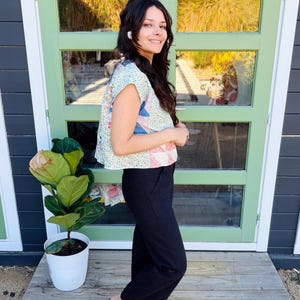 May include: A patchwork print top with a light blue background and a variety of colorful patterns. The top is paired with black pants and brown leather slip-on shoes. A potted plant is to the left of the person.
