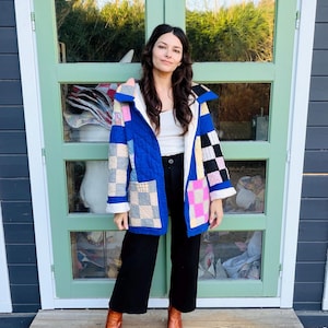 May include: A long-sleeved quilted jacket featuring a patchwork design with blue, pink, white, and grey squares. The jacket has a wide collar, two front pockets, and a white interior lining. It is worn over a white tank top and black trousers.