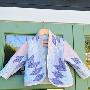 May include: A quilted jacket with a light pink and white floral pattern. The jacket features a geometric design with blue and pink floral accents. The sleeves and shoulders are solid light pink. The jacket is hanging on a wooden hanger.