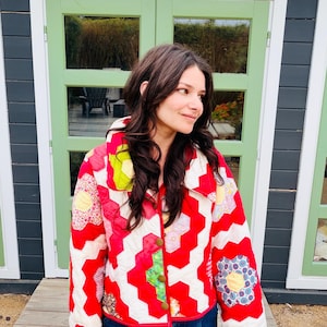 Puede incluir: Chaqueta acolchada con un patrón geométrico rojo y blanco. La chaqueta presenta un cuello blanco, botones dorados y parches florales coloridos. La persona lleva vaqueros azules. El fondo incluye puertas verdes y una terraza de madera.