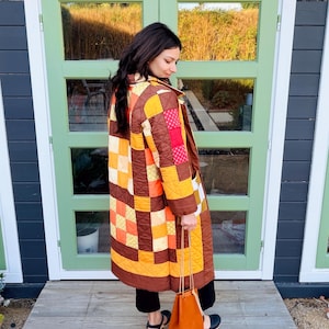 Pode incluir: Um casaco acolchoado de patchwork em tons de castanho, laranja e amarelo. O casaco apresenta um padrão geométrico e uma gola castanha. Uma pequena bolsa de couro laranja com cordão é segurada na mão.