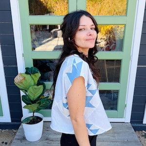 May include: A white top with blue geometric shapes. The top has short sleeves and a loose fit. The person is wearing black pants. A potted plant is to the left of the person. The background includes a green door and a dark gray wall.
