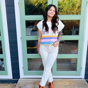 May include: A short-sleeved quilted top featuring a white base with a multi-colored star pattern and horizontal stripes in yellow, pink, and blue. The top is paired with white denim jeans and brown leather ankle boots with a pointed toe.