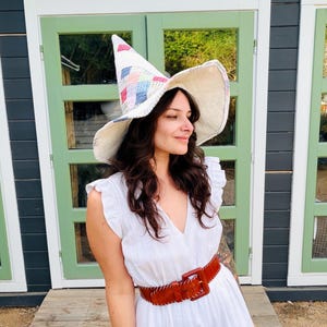 May include: A white sleeveless dress with ruffled shoulders and a deep V-neckline, paired with a wide brown leather belt featuring a large square buckle and visible stitching. A tall, pointed hat with a patchwork pattern in pastel colors.