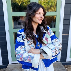 May include: A long-sleeved jacket constructed from a patchwork quilt featuring a mix of white, blue, and floral patterned fabric squares. The jacket includes a solid blue collar, blue trim along the front opening, and gold-toned metal snap buttons.