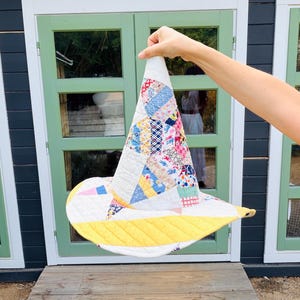 May include: A conical hat constructed from quilted fabric featuring a patchwork design of various floral and geometric patterns in shades of blue, pink, red, and white, finished with a solid yellow quilted brim and white quilted accents.