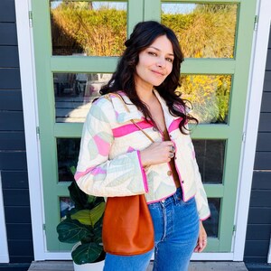 May include: A quilted jacket with a cream background, pink and green geometric patterns, and pink trim. The jacket is paired with blue jeans and a brown leather drawstring bag. The background features a green door and a house.