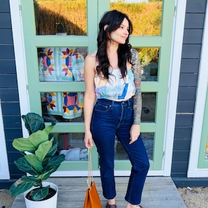 Könnte beinhalten: Ein Patchwork-Crop-Top in Pastellfarben, kombiniert mit dunkelblauen Jeans und braunen Clogs. Eine kleine, orangefarbene Leder-Kordelzugtasche wird gehalten. Links befindet sich eine Topfpflanze.