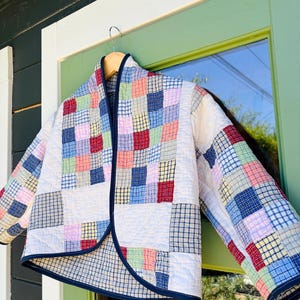 May include: A quilted jacket featuring a patchwork design of various plaid and checkered fabric squares in shades of red, blue, pink, green, and white. The garment has long sleeves, a rounded open front, and is finished with a dark blue trim.