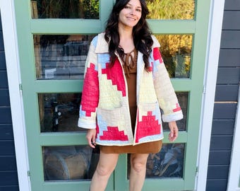 May include: A quilted jacket featuring a geometric patchwork pattern in cream, red, and light blue. The jacket has a collar, long sleeves, and two front pockets. Underneath is a brown dress with a tie-front neckline and green slip-on shoes.