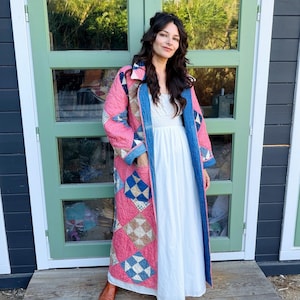 May include: A long, quilted coat featuring a patchwork design of pink, blue, and tan fabric squares. The coat has a collar, long sleeves, and two front pockets. Underneath, a floor-length white dress with a smocked bodice is worn with brown boots.