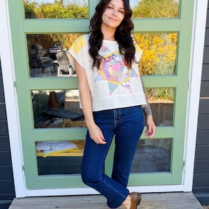 Puede incluir: Una blusa de manga corta color crema con un diseño de estrella de patchwork multicolor en la parte delantera, combinada con jeans azul oscuro y zuecos de cuero marrón. La blusa tiene detalles amarillos en los hombros y un corte holgado.