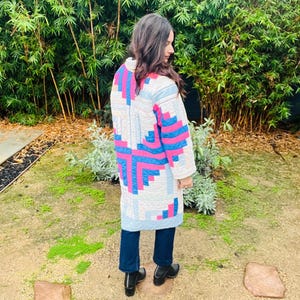 Pode incluir: Um casaco longo acolchoado com um padrão geométrico em tons de azul, rosa e branco. O casaco é usado sobre jeans azul escuro e botas pretas. O fundo apresenta bambu verde exuberante.