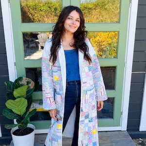 Könnte beinhalten: Ein langer, vorne offener Mantel aus einem Patchwork-Quilt mit einer Vielzahl kleiner, mehrfarbiger floraler und geometrischer Stoffquadrate in Blau-, Rosa-, Gelb- und Weißtönen, ausgestattet mit langen Ärmeln und zwei großen aufgesetzten Taschen.