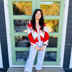 May include: A red and white patchwork jacket with floral accents is worn with white pants and brown shoes. The jacket has a bold design, and the outfit is completed with a white top. The person is standing in front of a green door.
