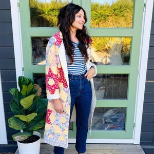 Könnte beinhalten: Ein langer, gesteppter Mantel mit einem Patchwork-Design aus roten, gelben und floralen Stoffmustern. Der Mantel wird über einem blau-weiß horizontal gestreiften T-Shirt und einer dunkelblauen Jeans getragen, kombiniert mit schwarzen Leder-Clogs.