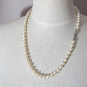 May include: A single strand necklace composed of uniform, round, off-white pearls. The necklace features a decorative, rectangular silver-toned metal clasp with intricate detailing, positioned on the right side of the strand.