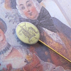 May include: A gold and silver oval pin with an engraved monogram. The pin has a long, thin, gold stem.