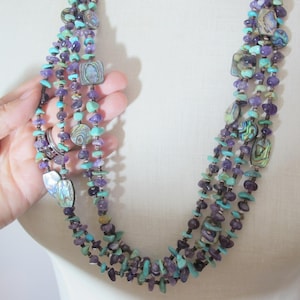 Puede incluir: Un collar de múltiples hilos que presenta una combinación de pequeñas astillas de amatista púrpura irregular, cuentas de piedra turquesa brillante y acentos de concha de abulón iridiscente. Los hilos están dispuestos en un diseño en capas.