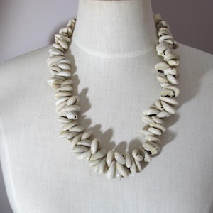 May include: A white shell necklace displayed on a mannequin. The necklace is made of numerous small, oblong shells strung together. The shells are a creamy white color, and the necklace is draped around the mannequin's neck.