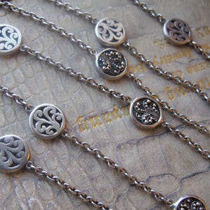 Può includere: Collane argentate con pendenti circolari e ornati. Ogni pendente presenta un design dettagliato e a spirale. Le catene sono delicate e le collane sono disposte diagonalmente, creando un motivo su uno sfondo strutturato.