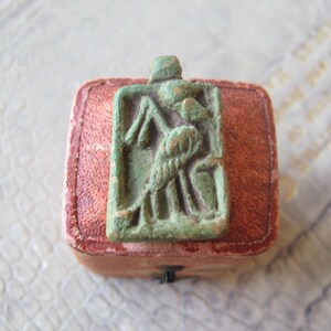 May include: A green rectangular pendant with an embossed figure design, possibly Egyptian, resting on a small, square, red leather box. The item appears to be a piece of jewellery or a collectible, presented in a vintage style.