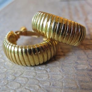 May include: A pair of gold-colored hoop earrings. The earrings have a ribbed design and a shiny finish. The earrings are displayed on a textured surface.