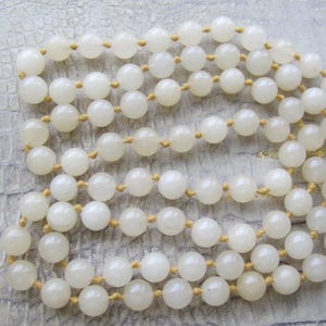 May include: A long necklace composed of numerous round, off-white beads. The beads are strung together with a light-colored cord, creating multiple strands. The necklace is displayed on a textured, light gray surface.