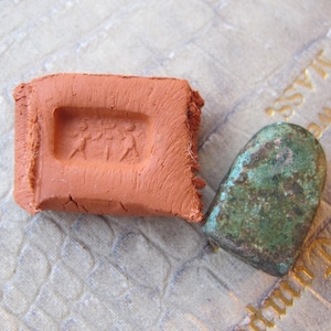 Può includere: Un mattone color terracotta con un disegno in rilievo di tre figure, accanto a un piccolo oggetto metallico arrotondato e con patina verde. Gli oggetti poggiano su una superficie strutturata di colore chiaro.