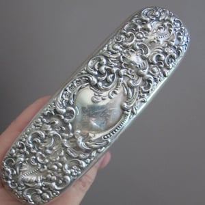 May include: An elongated, oval-shaped silver case featuring intricate, raised scrollwork and floral repoussé detailing across its entire surface. The center of the object displays a smooth, polished, unadorned shield-shaped cartouche.