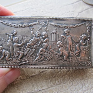 May include: A silver rectangular box with an ornate design featuring cherubs playing music and holding scrolls. The box has a beaded border and a raised design.