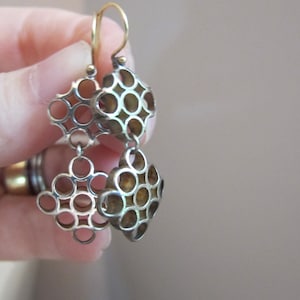 May include: Silver-toned dangle earrings with a geometric openwork design. Each earring features two diamond-shaped sections connected by small rings. The earrings have gold-colored ear wires and a light brown background.