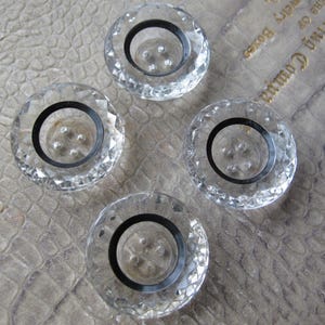 May include: Four clear, faceted glass buttons with a black ring around the center. The buttons have a circular shape and a decorative, geometric design. These buttons are likely for sewing or crafts.